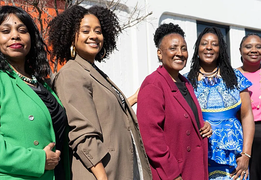 Five women standing outside, smiling in colorful professional outfits.