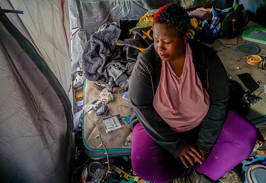 Woman sits inside a cluttered tent, looking down, surrounded by personal belongings.