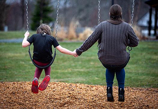 A child and an adult sit on swings holding hands, seen from behind, in a grassy playground with wood chips on the ground.
