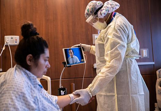 A hospital worker speaks through a remote Spanish translator while preparing a COVID-19 patient take her first steps after being