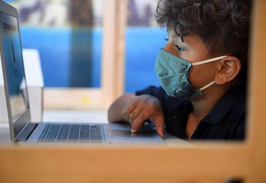 A student follows along remotely with their regular school teacher's online live lesson from a desk separated from others by pla