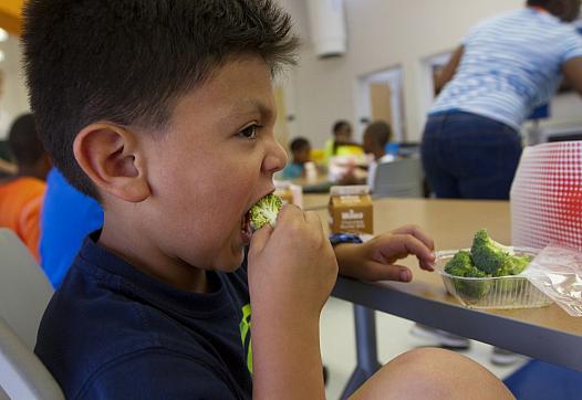 Six-year-old Jacob Portillo says he likes eating his broccoli at summer camp at Park View Recreation Center in Washington, D.C. The camp offers free lunches — funded by the USDA — to all participants and children in the community. For some, the meal fills a nutritional gap that’s crucial during the summer months when kids are out of school. Photo by Margaret Myers/NewsHour