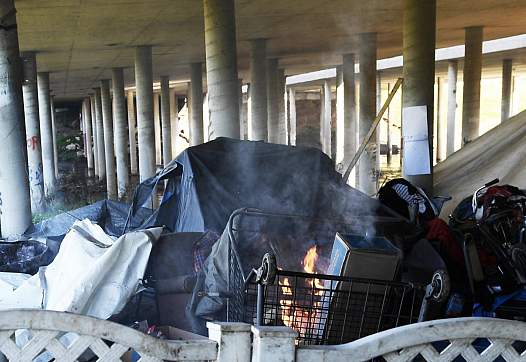 Homeless people wrap themselves in tarps as they try to keep warm near a fire at a homeless encampment beneath the El Dorado St